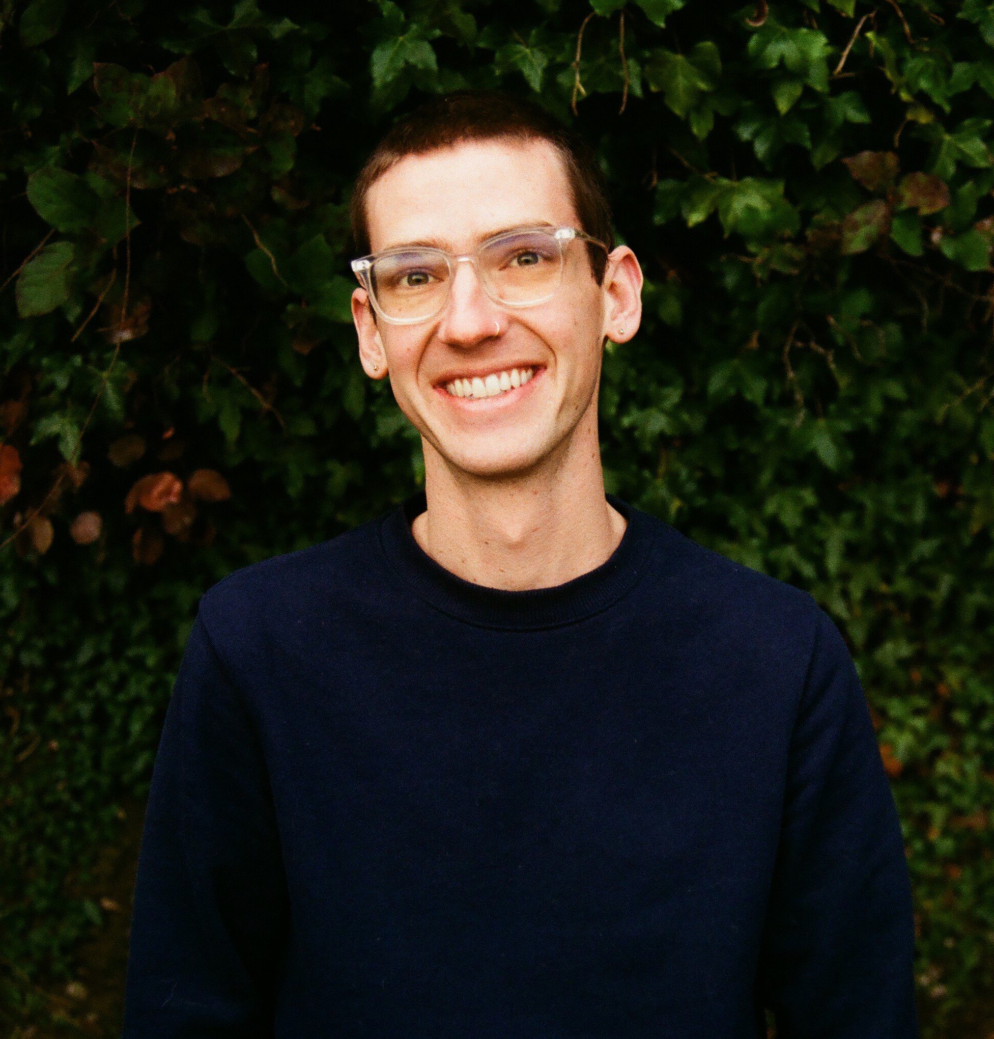 Kevin Steen's headshot. Kevin smiles in front of a wall of leaves. Kevin wears clear glasses, a dark long sleeved shirt, and short cropped brown hair.