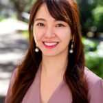 Lauren Kang's headshot. Lauren poses outside in front of green trees. Smiling, Lauren wears a taupe blouse and long reddish-brown hair partly pulled back.