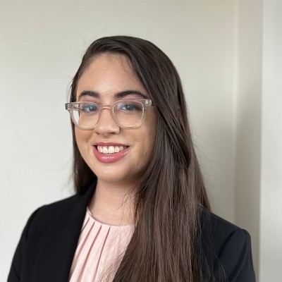 Adriana Herrera's headshot. Adriana smiles in front of a white wall, wearing a light pink top and a black blazer. Adriana has long straight dark brown hair and clear glasses.