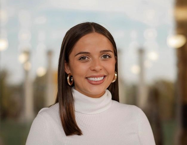 Mackenzie Gettel's headshot. Mackenzie smiles in front of a blurred background of columns and buildings. Mackenzie wears a white turtleneck and has medium length brown straight hair.