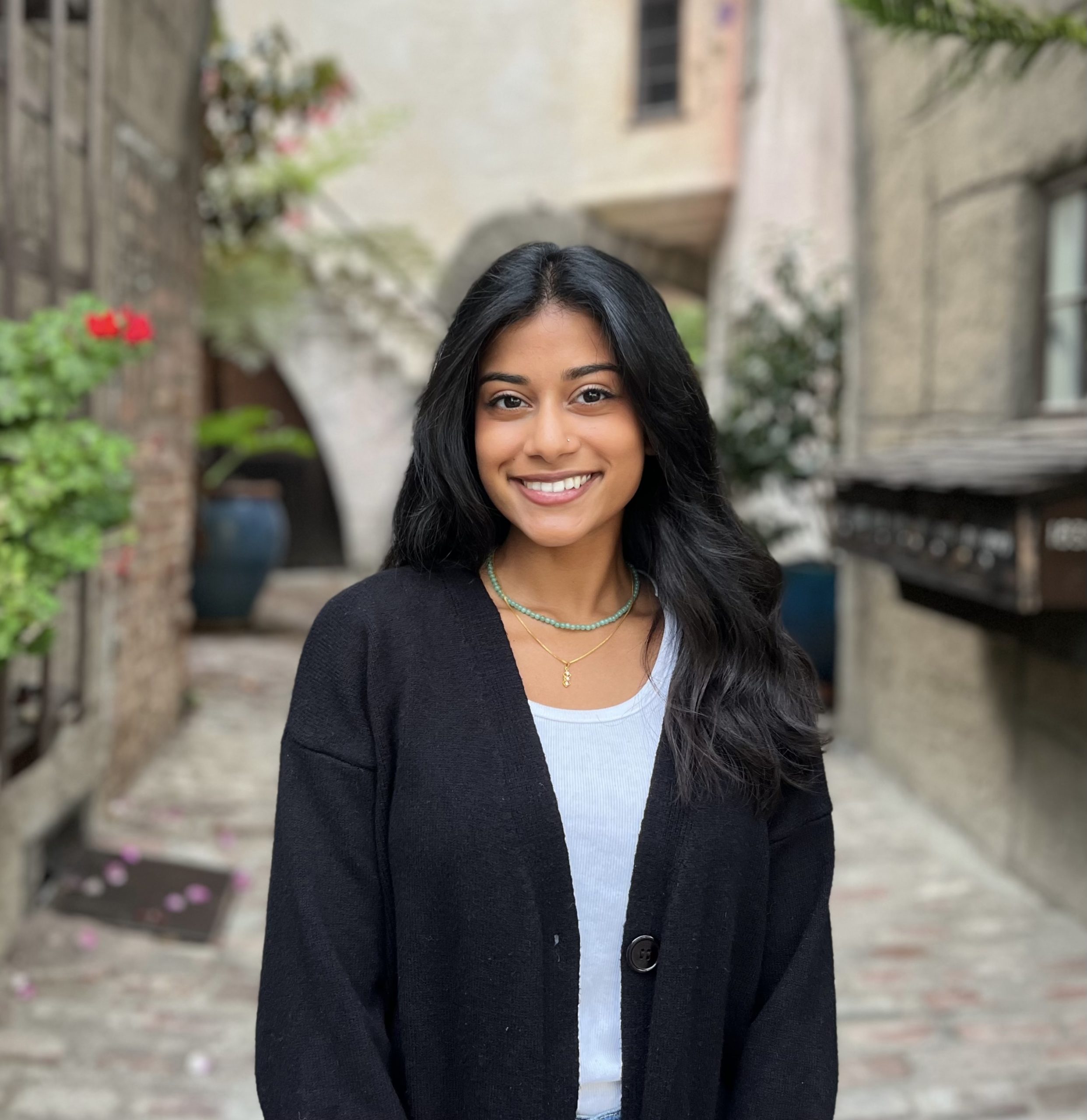 Bhavya Sukhavasi's headshot. Bhavya is standing in a cobbled alleyway between stone buildings. Smiling, Bhavya wears a white shirt and a black cardigan, with long black wavy hair.
