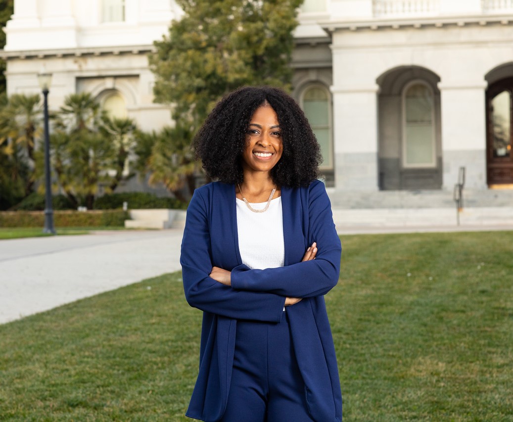 Tiffaney Boyd's headshot. Tiffaney poses in green courtyard in front of a white building. Arms crossed and smiling, Tiffaney wears a white blouse and a navy suit. Tiffaney has medium-length black curly hair.