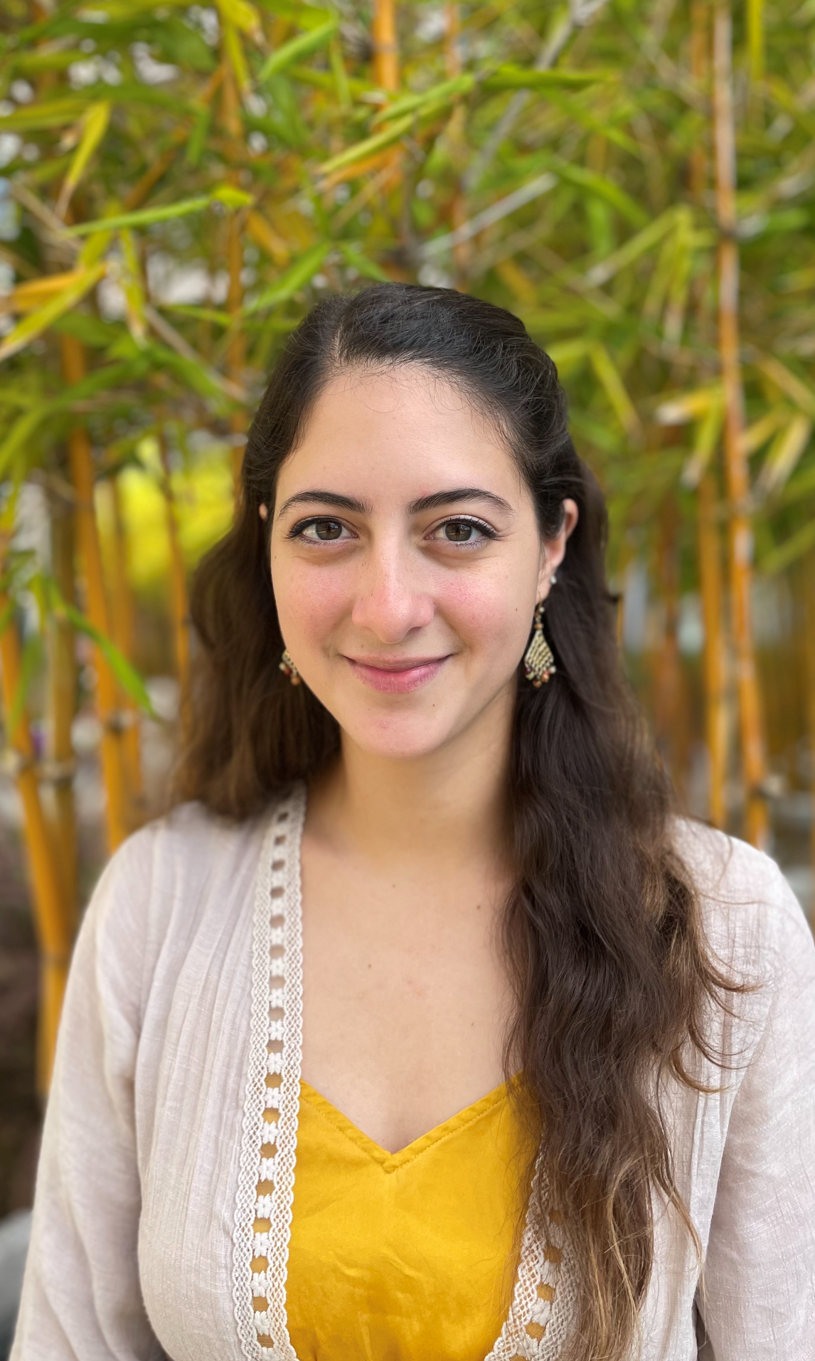 Marian Avila Breach's headshot. Marian is slightly smiling standing in front of bamboo trees with green leaves. Marian is wearing a yellow blouse with a white overshirt, with long wavy brown hair.