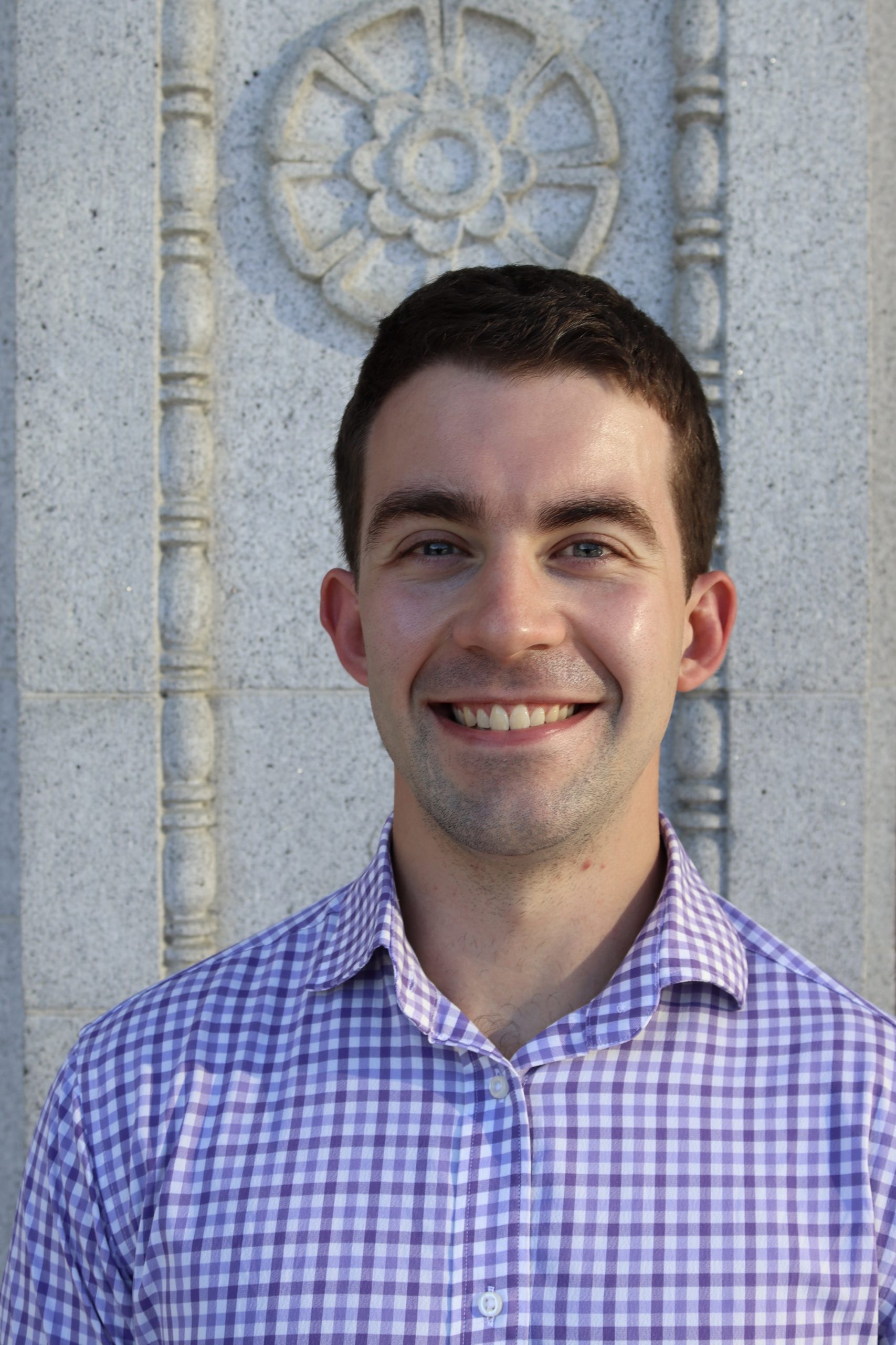 Peter Mason's headshot. Peter is smiling standing in front of a decorative stone structure. Peter is wearing a blue and white collared shirt and has short brown hair.
