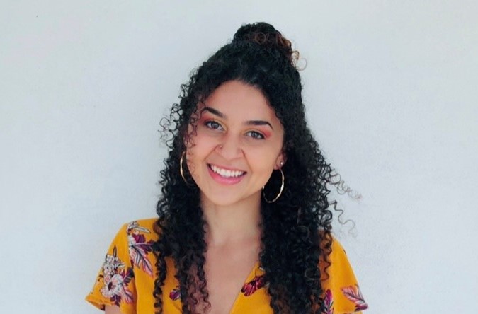 Julianna Gay's headshot. Julianna smiles in front of a light blue background. Julianna wears a floral yellow top and has long black curly hair.