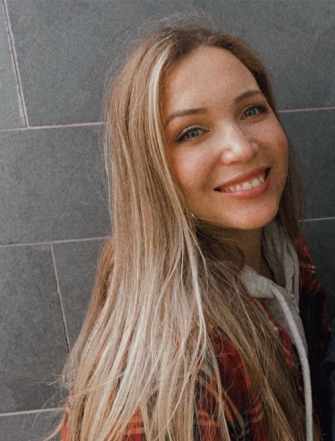 MacKenna Alvarez's headshot. MacKenna is smiling against a gray stone wall, wearing a red plaid flannel jacket and a gray sweatshirt. MacKenna has long straight blonde hair.