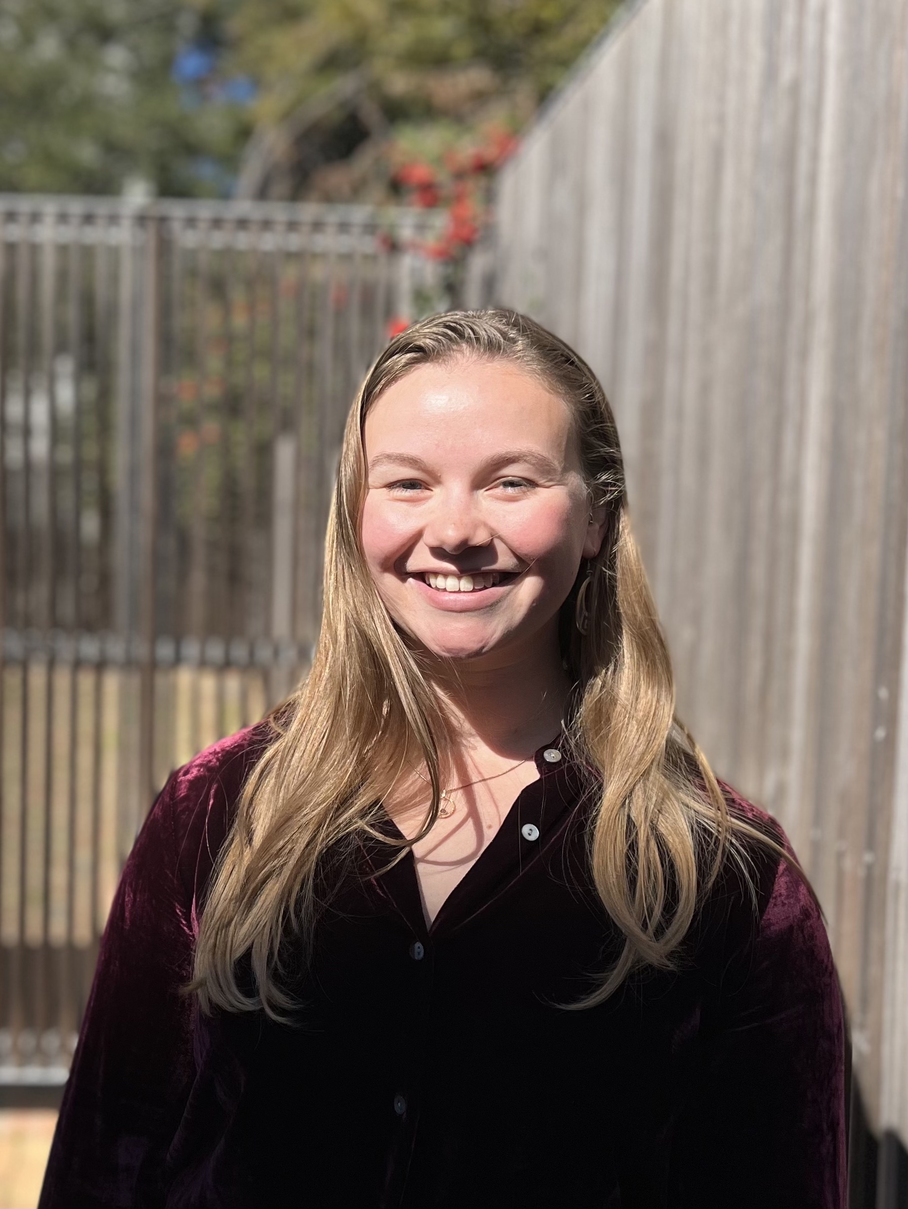 Nora Ellmann's headshot. Nora is smiling near a wooden fence. Wearing a maroon velvet button up shirt, Nora has long wavy dark blonde hair.
