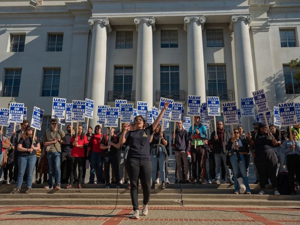 Students stand in front of a university building on UC Berkeley\'s campus. One person in the middle speaks through a megaphone. The students carry signs reading \"UAW ON STRIKE: UNFAIR LABOR PRACTICE.\"