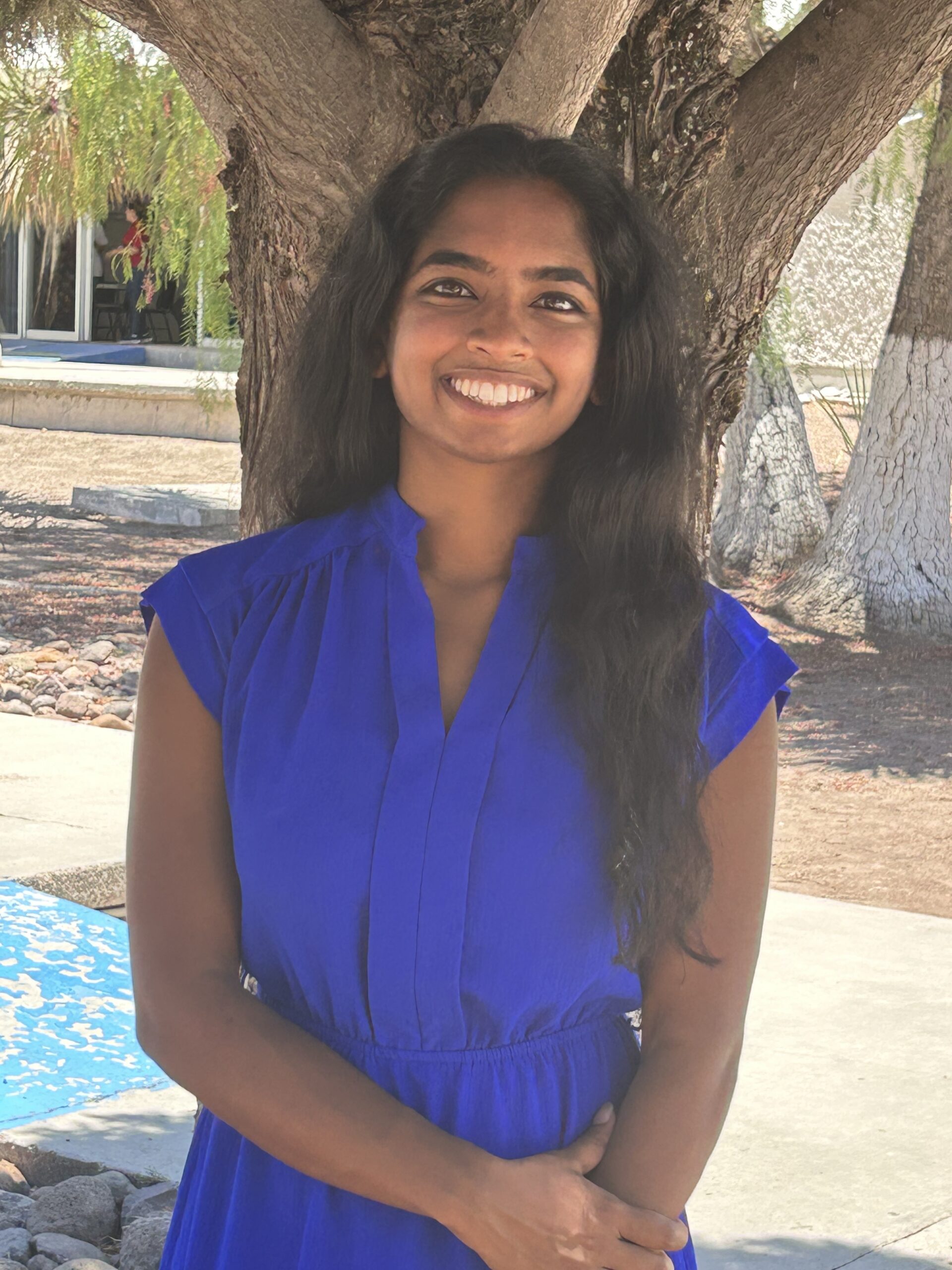 Meghana Ravikumar's headshot. Meghana smiles a big smile in front of a tree and courtyard. She is wearing a royal blue dress and has long wavy black hair.