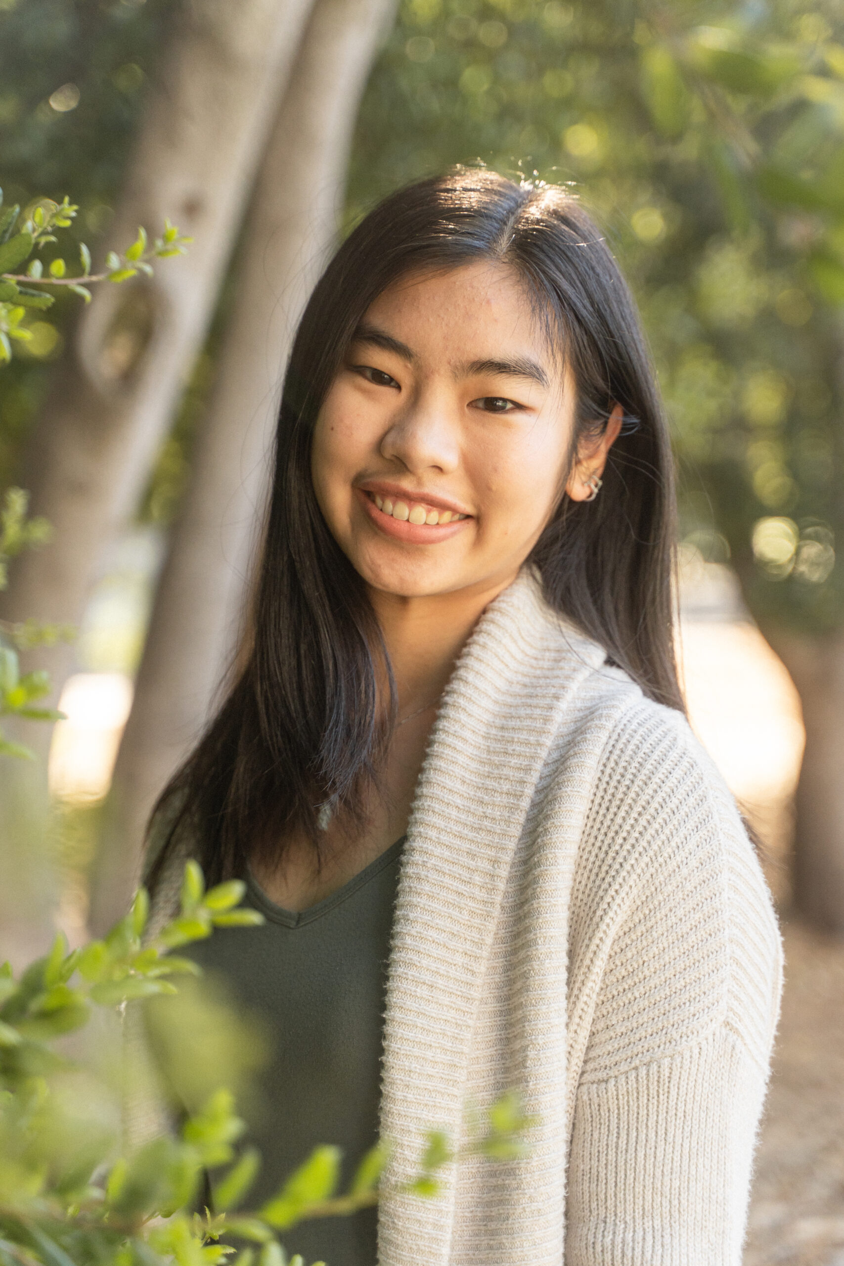Ianna Zhu's headshot. Ianna poses outdoors with some leaves in front of her and a large tree behind her. She wears a cream-colored cardigan over a green shirt and has long straight black hair.