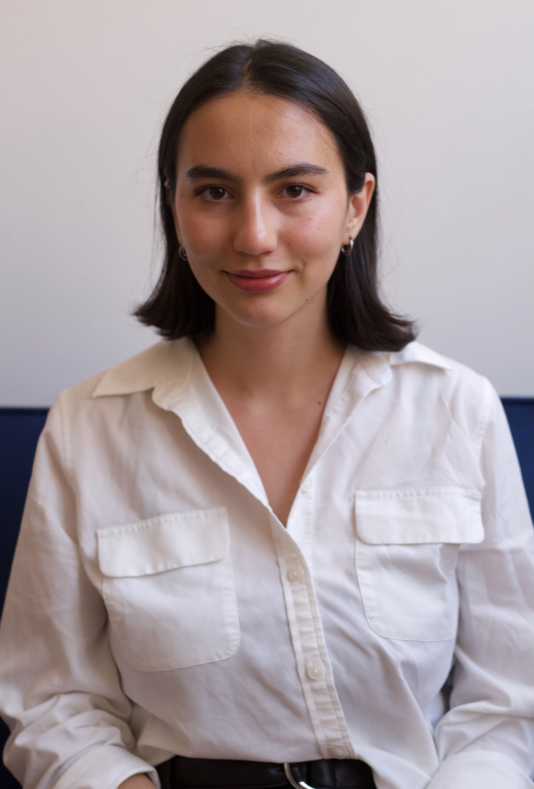 Samahria Alpern's headshot. Samahria is smiling slightly against a white wall. She has shoulder-length dark brown hair and is wearing a white button-up shirt. 