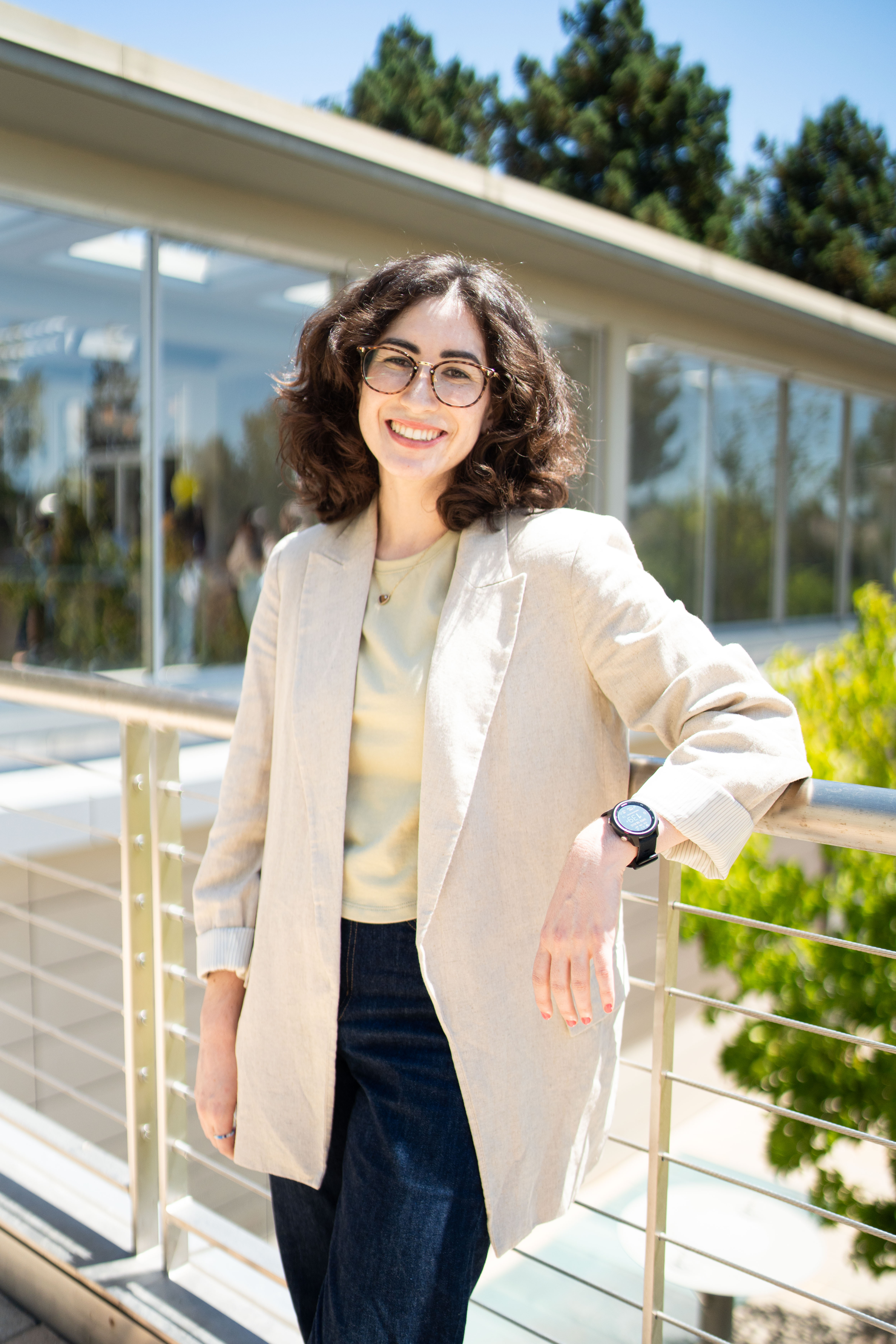 Lande Watson's headshot. Lande grins while surrounded by flowers and plants. She wears a black shirt, a green button-up, and glasses, and has shoulder-length curly brown hair.