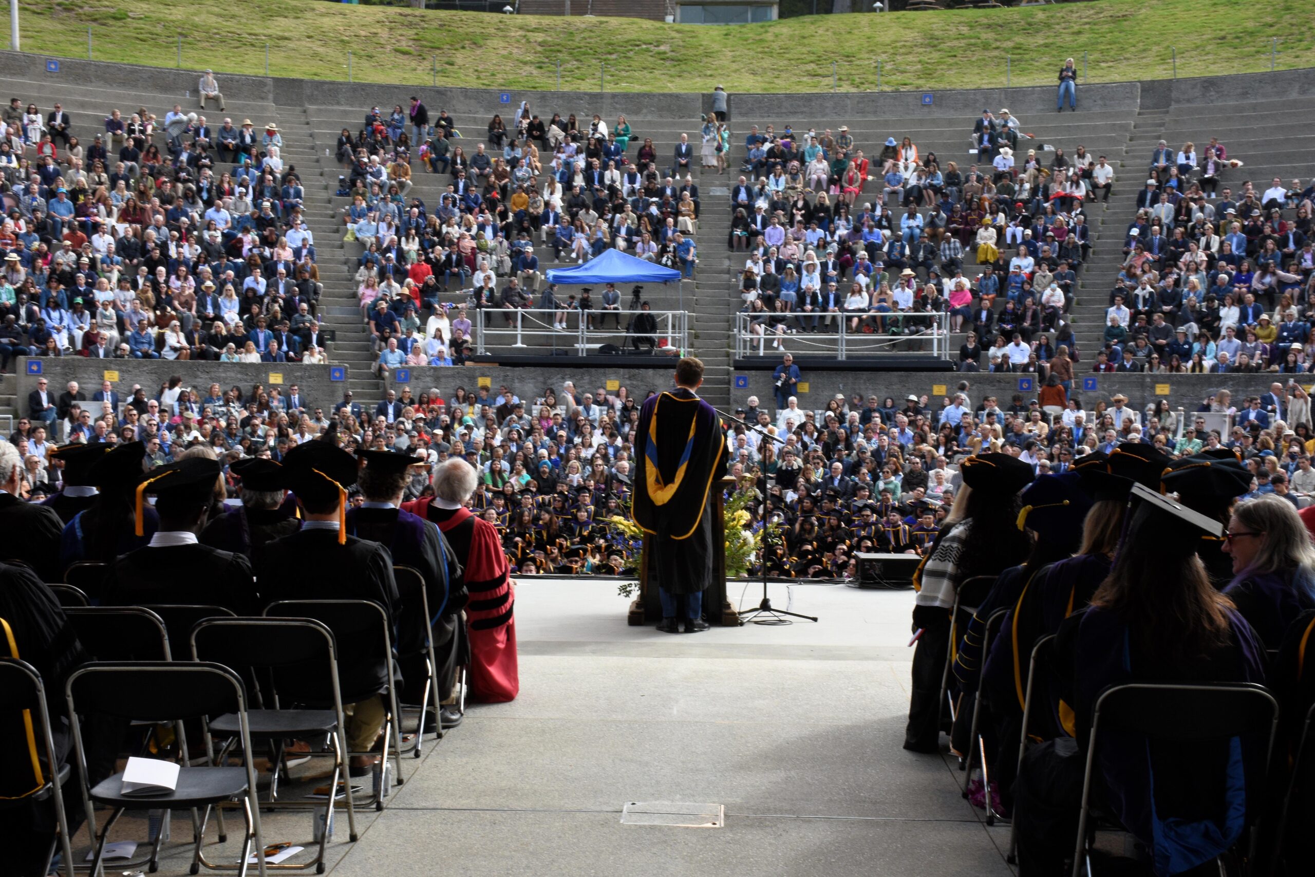 View of Greek Theatre from stage out to audience with speaker at podium and faculty seated on stage in regalia.