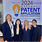Four people posing in front of Patent sign
