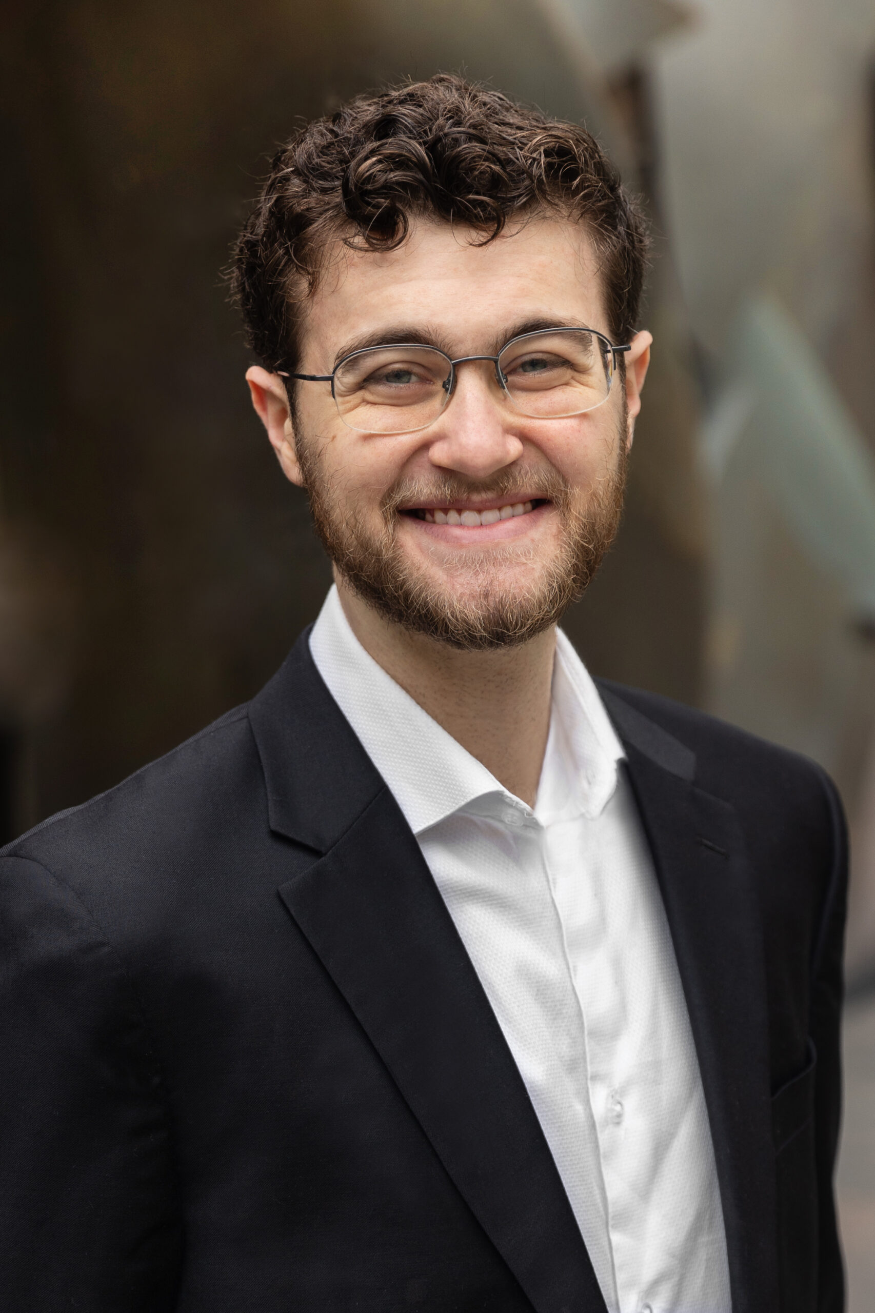 Student Adam Aronovsky smiles in front of a blurred background, wearing a black suit jacket and white button up shirt. He wears glasses, has a short beard, and short curly brown hair.