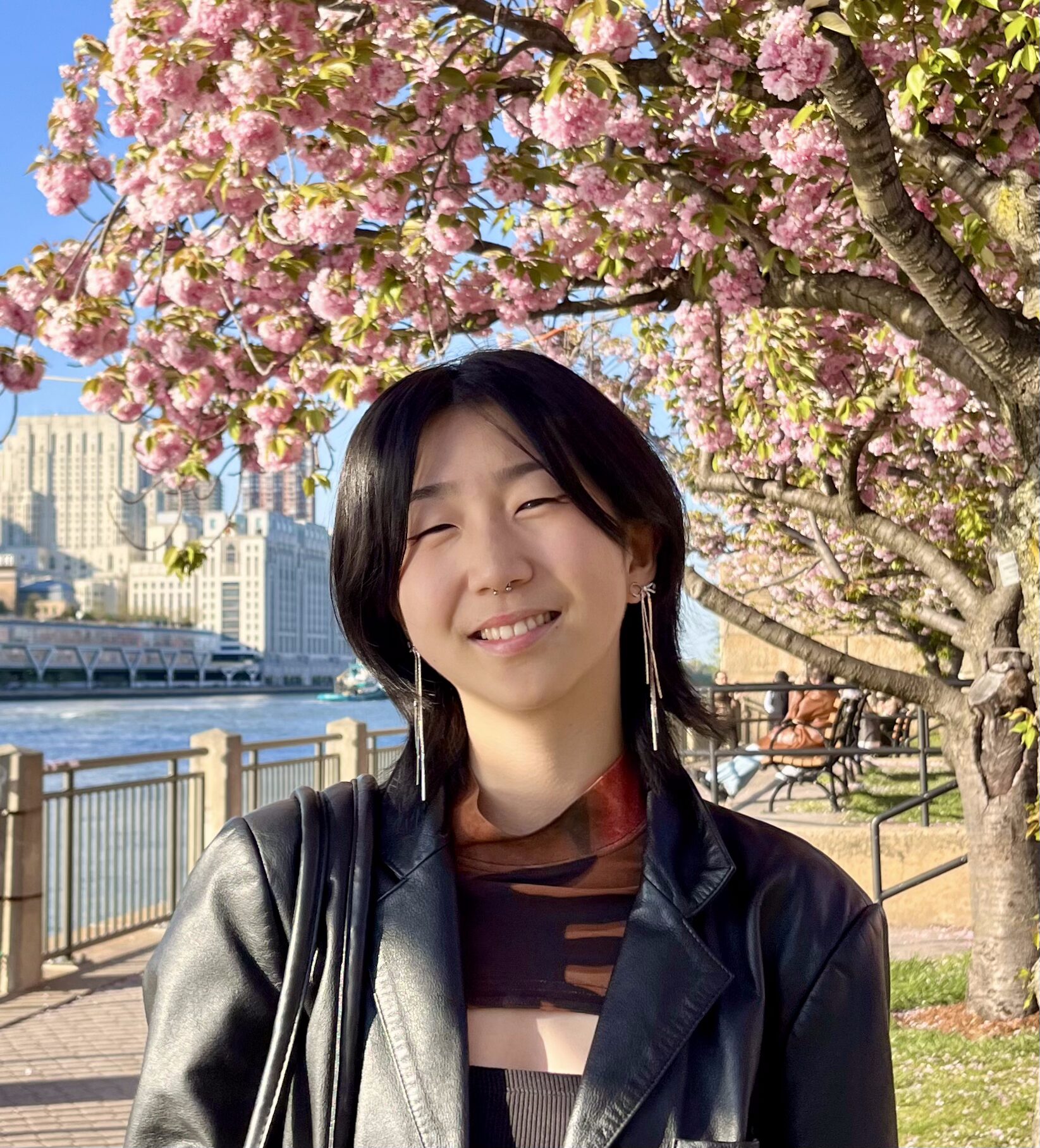 Student Sunnie Liu smiles in front of a blossoming pink tree and a waterfront. They are wearing a black leather jacket, orange and black shirt with cut outs, and have medium length black hair.