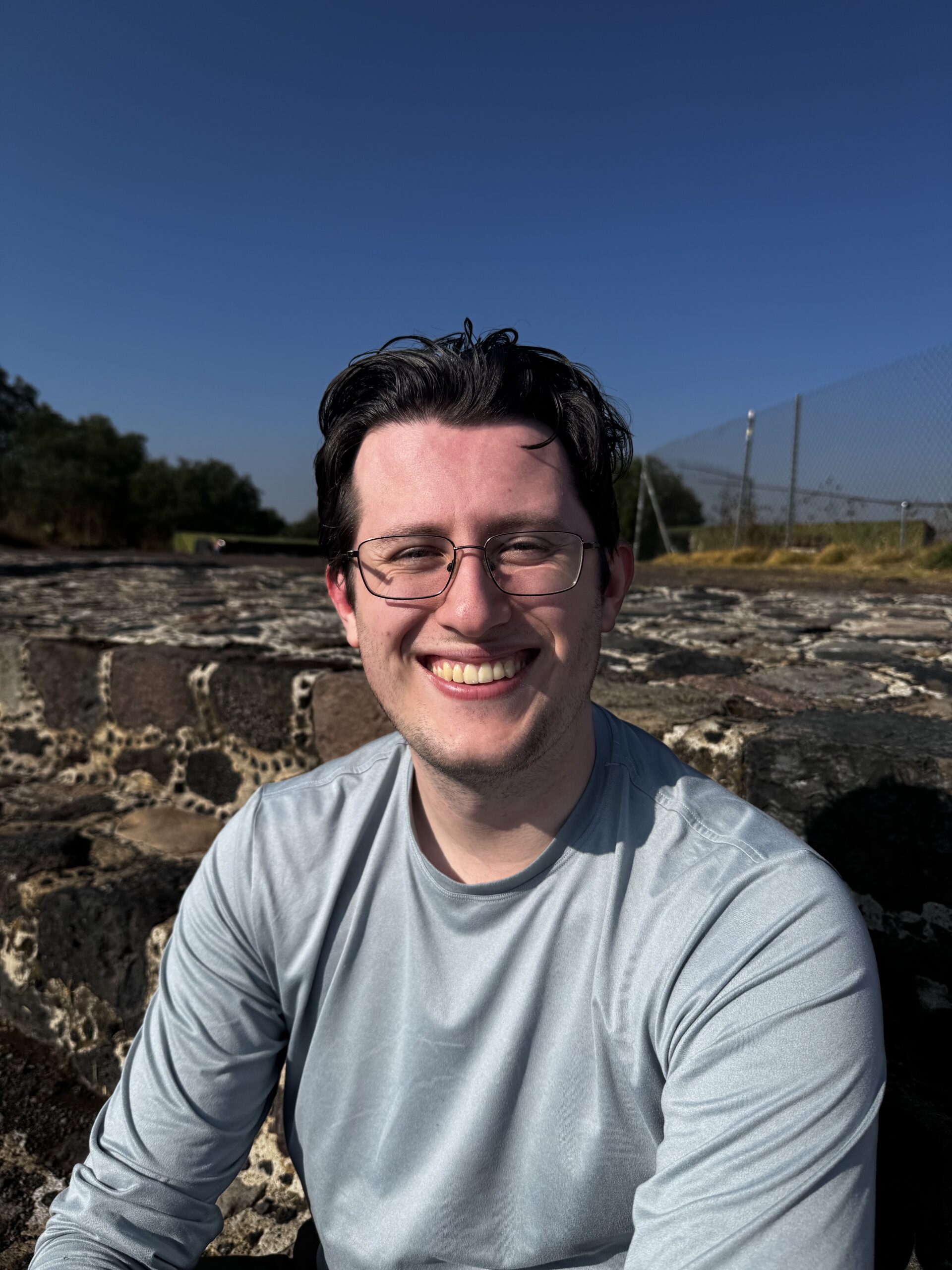 Student Quinn Samouilidis smiles, sitting on stairs with decorative mosaic stones. He's wearing a long sleeve gray shirt and glasses, and has short dark brown hair.
